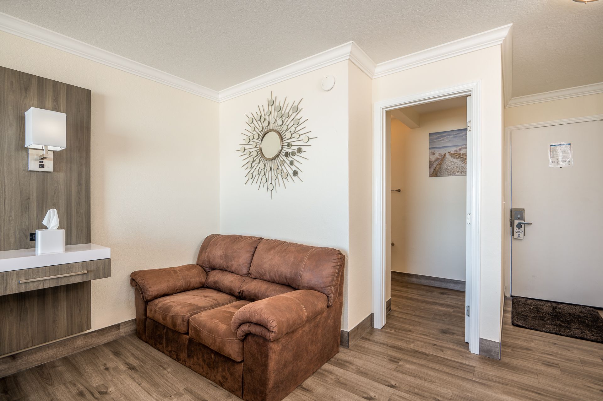 A hotel room corner featuring a brown loveseat, a floating wooden desk, a white wall-mounted lamp, and a doorway.