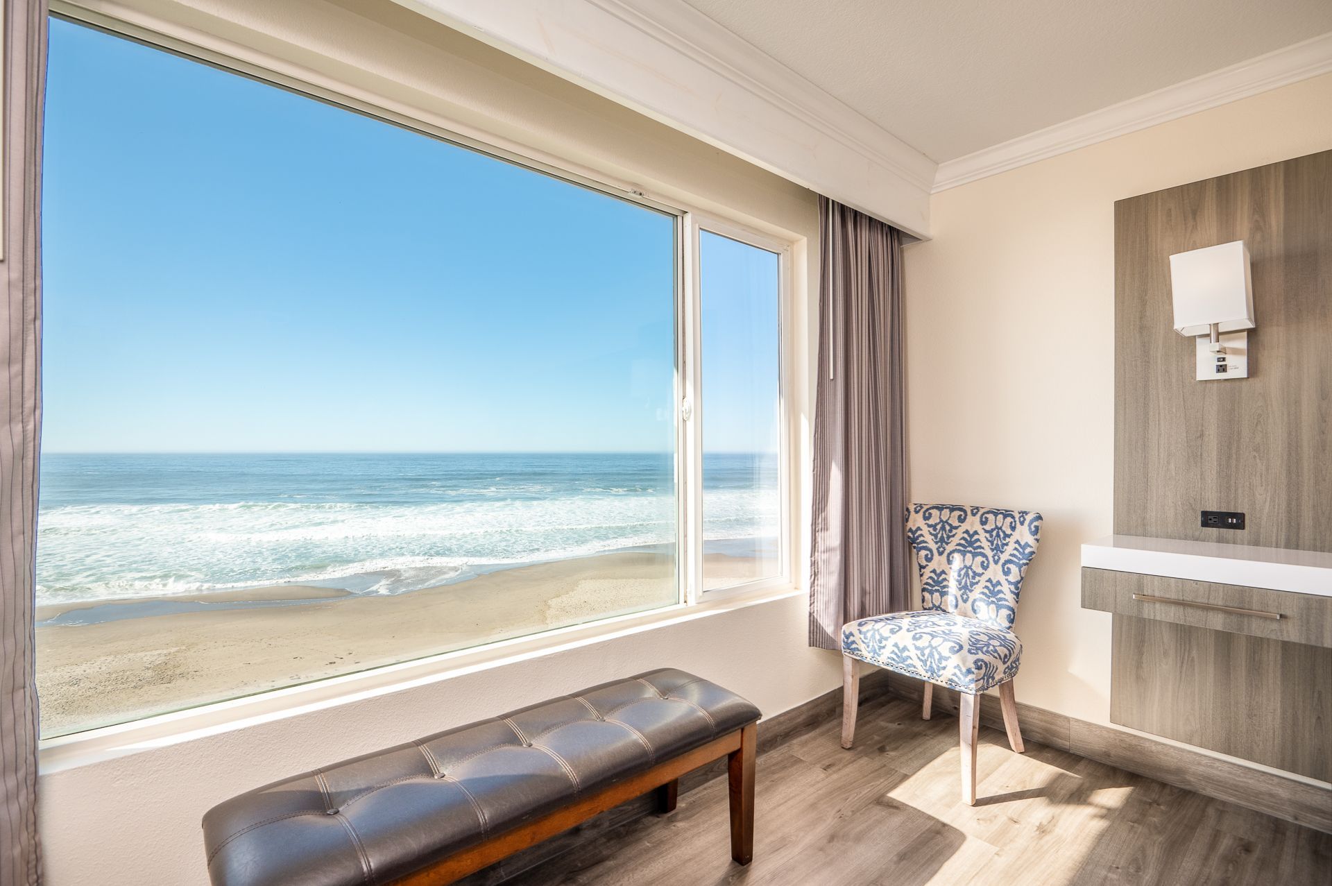 A hotel room with a large window overlooking the ocean, featuring a brown leather bench and a patterned chair.