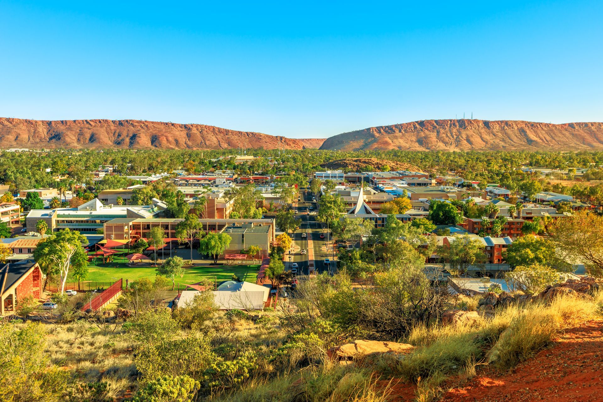 A high-angle view of Alice Springs, Australia, featuring a dense town center nestled below prominent red desert ranges.