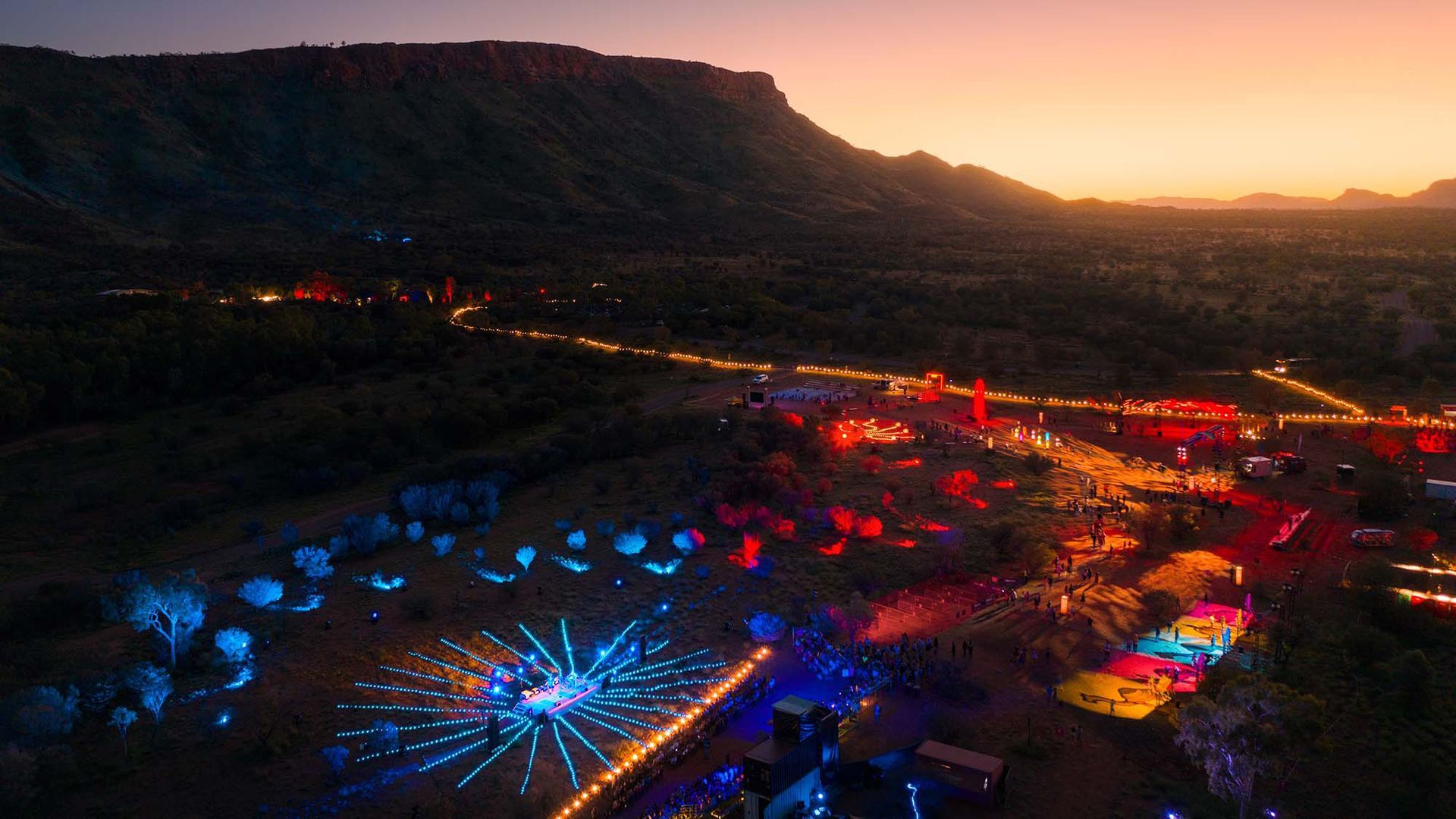 Aerial view of a vibrant desert music festival at sunset, with illuminated art installations and glowing paths.