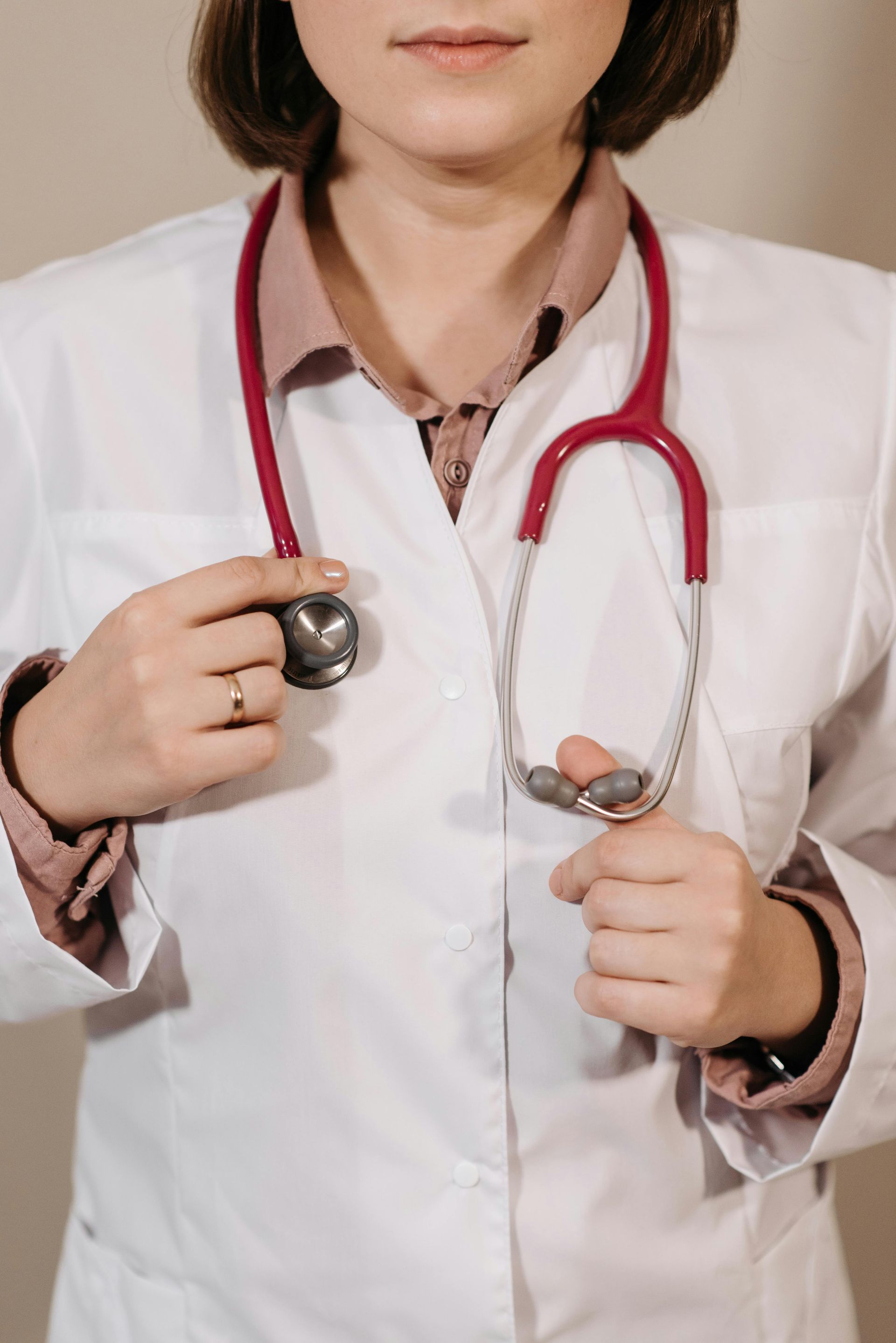 A person in a white lab coat with a red stethoscope draped around their neck, holding the chest piece.
