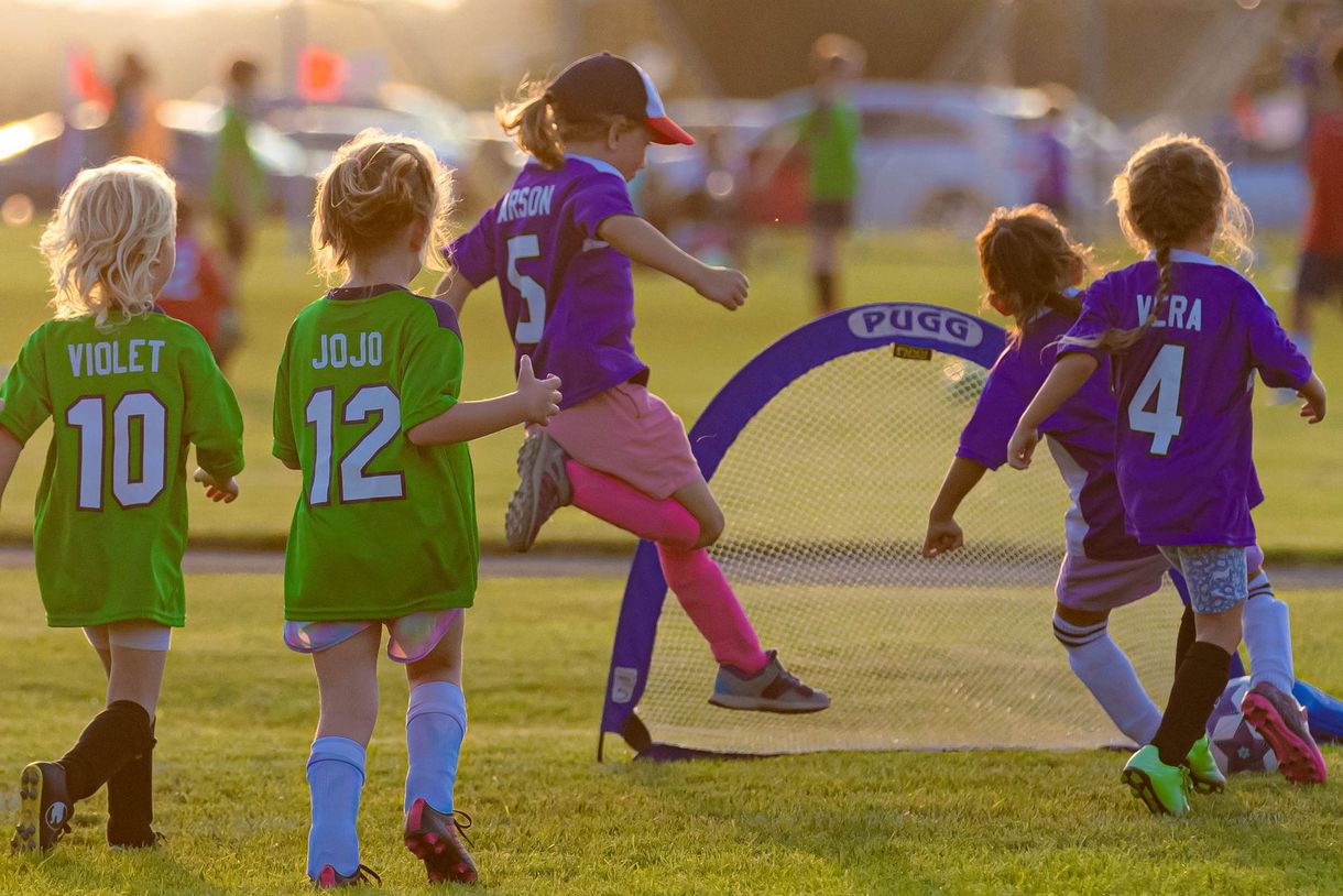Children playing soccer outdoors; girl in purple jersey kicks the ball toward a small net.
