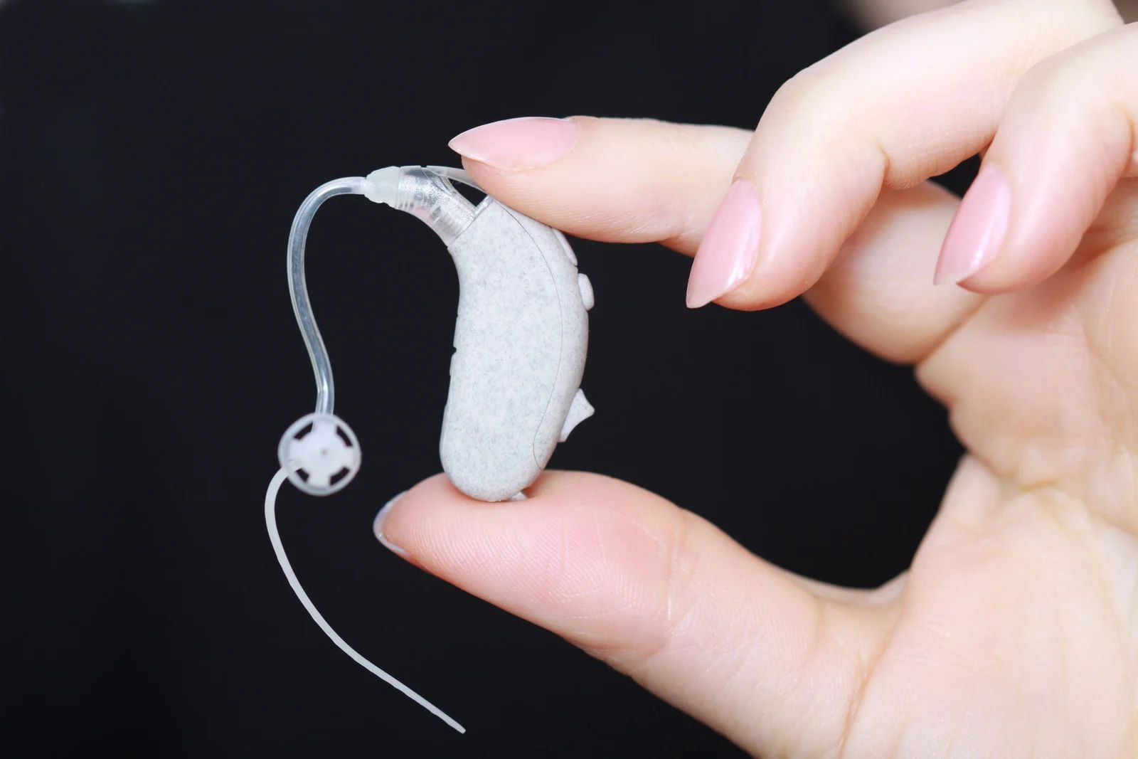 Hand holding a beige hearing aid with a clear tube and small earpiece against a black background.