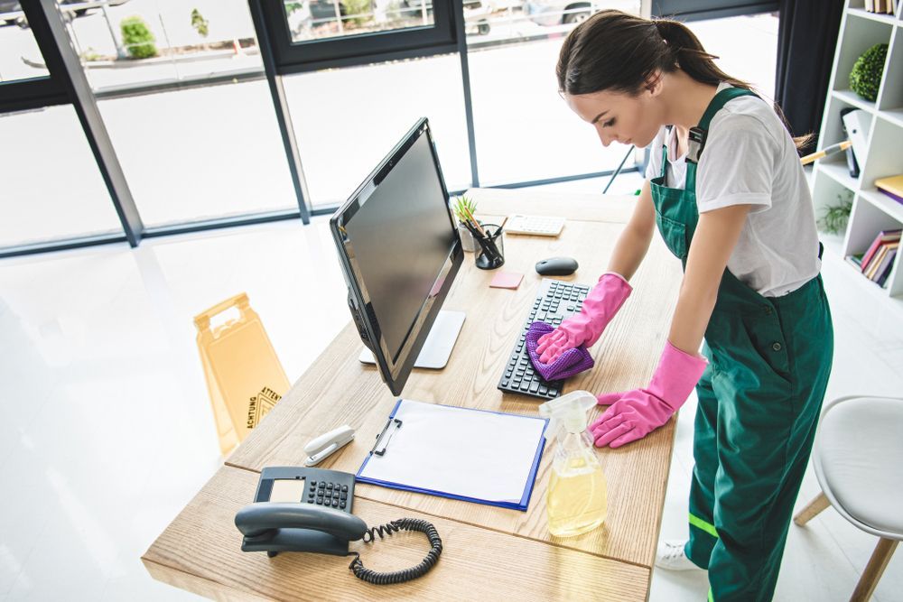 A woman is cleaning an office desk with a computer and a phone.