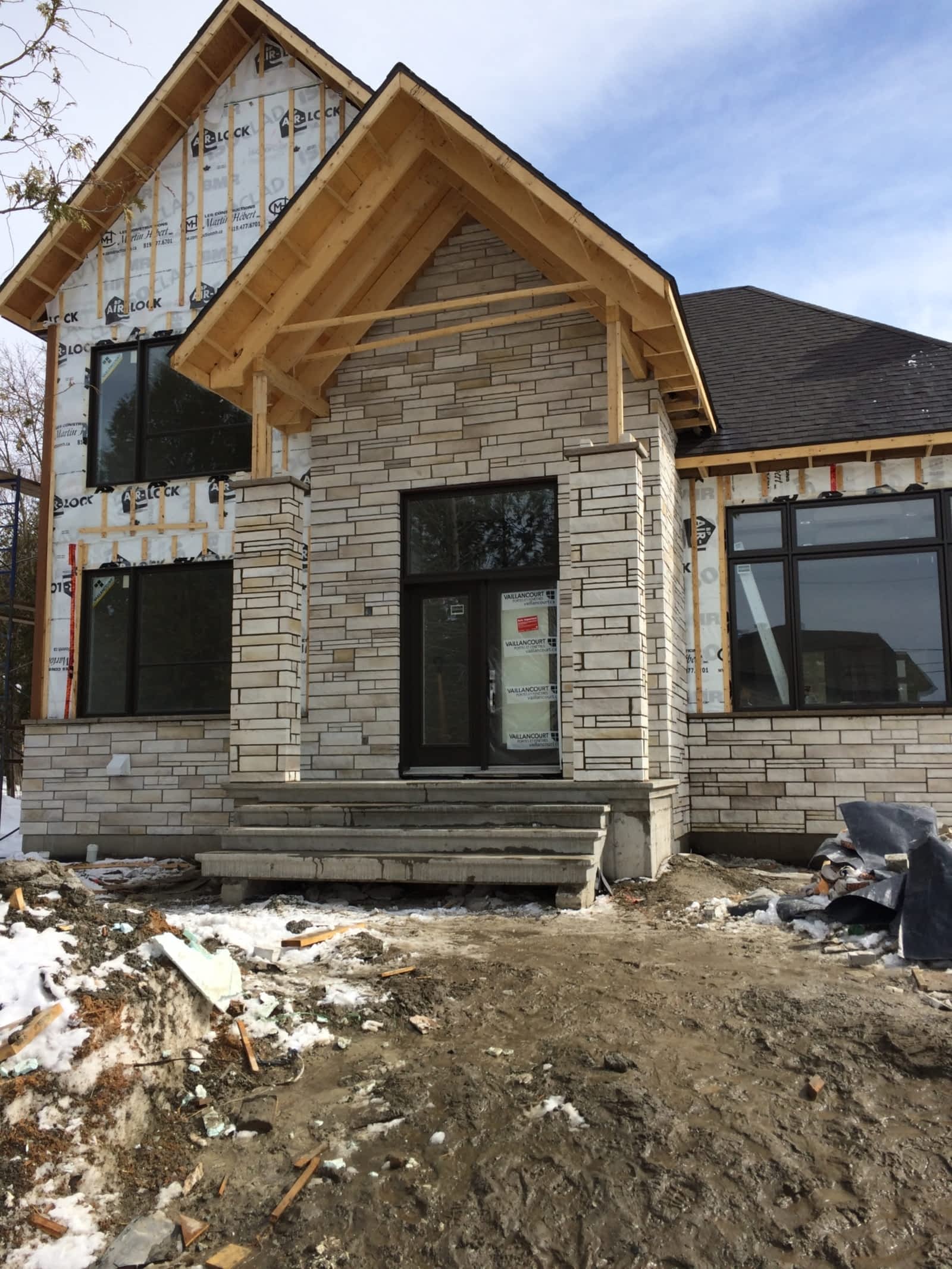 Extérieur de la maison en construction, façade en pierre, porche d'entrée, fenêtres à cadre sombre, toiture partiellement installée, neige au sol.