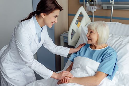 A medical professional in a white coat smiles while holding the hand of a patient resting in a hospital bed.