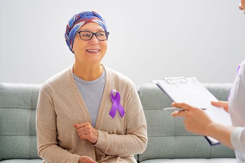 A person wearing a purple awareness ribbon on a beige cardigan sits on a couch while talking to a healthcare provider.