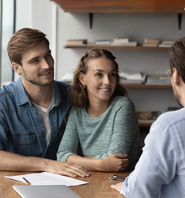 A couple sits at a wooden table in an office, smiling while listening to a professional sitting across from them.