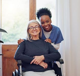 A smiling caregiver in blue scrubs stands behind a person in a wheelchair