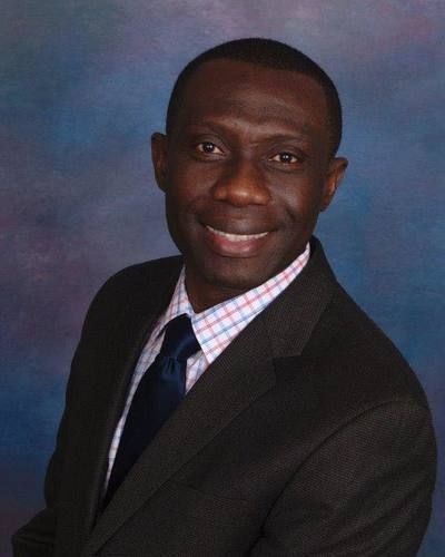 A person wearing a dark suit, a light-colored plaid shirt, and a dark blue tie, smiling against a blue mottled background.