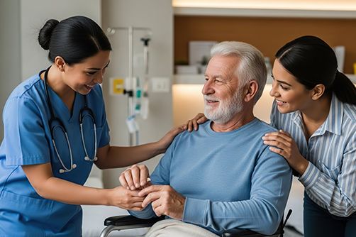 A medical professional in blue scrubs and a person in a striped shirt support an older individual sitting in a chair.