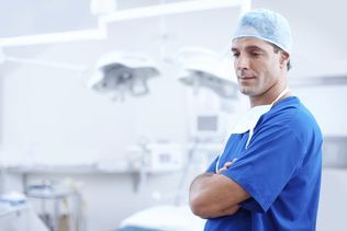 A surgeon in blue scrubs and a cap standing with arms crossed in a brightly lit, sterile operating room.