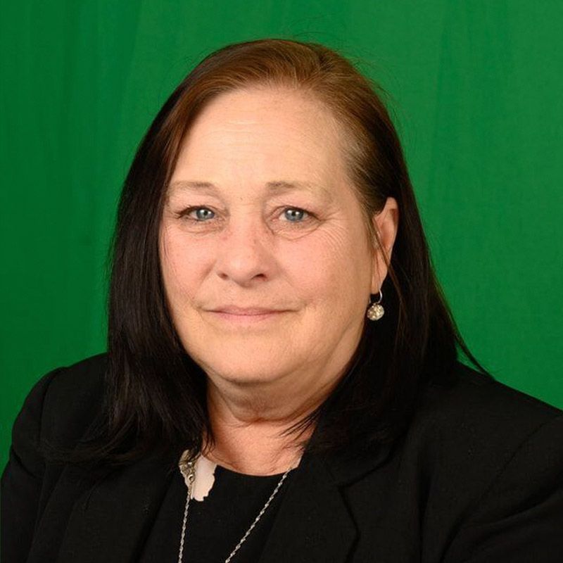 A professional headshot of a woman wearing a black blazer and a necklace, set against a solid green background.
