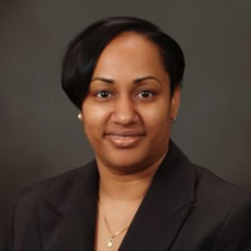 A professional headshot of a person wearing a black blazer and a delicate necklace against a plain dark background.