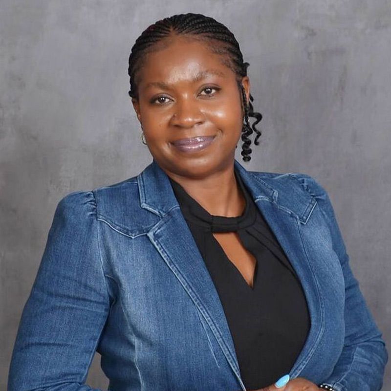 A person wearing a denim blazer over a black top, posing against a gray background.