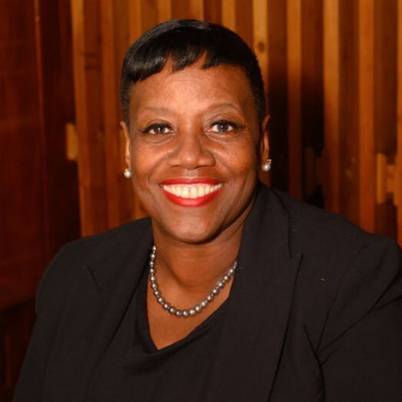 A smiling person with short dark hair, wearing a black blazer and a pearl necklace, stands against a wooden paneled wall.