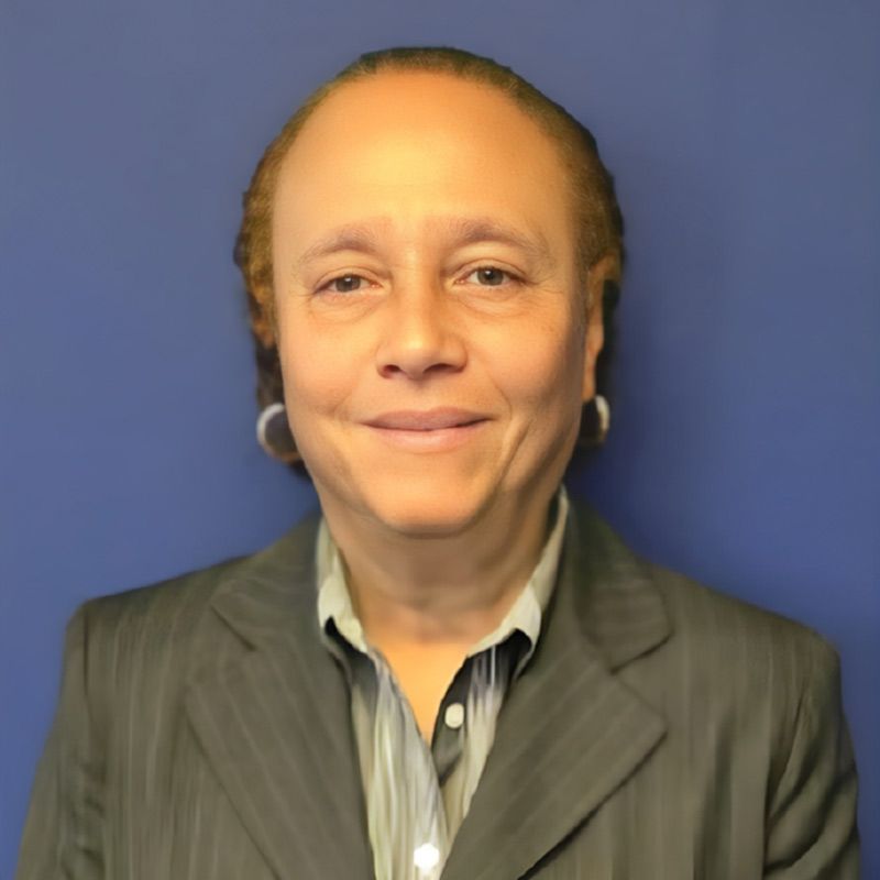 A smiling person wearing a pinstripe blazer and collared shirt stands against a solid blue background.