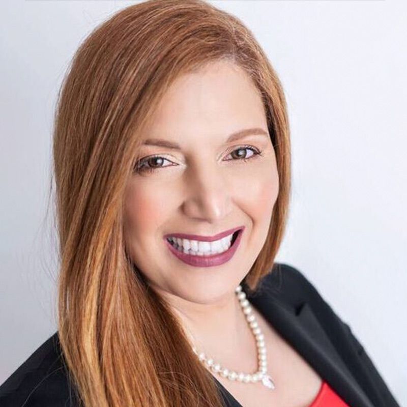 A smiling professional with shoulder-length auburn hair, wearing a black blazer, red top, and a pearl necklace.