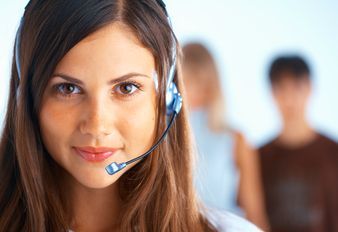 A customer service representative wearing a headset smiles warmly, with blurred colleagues visible in the background.