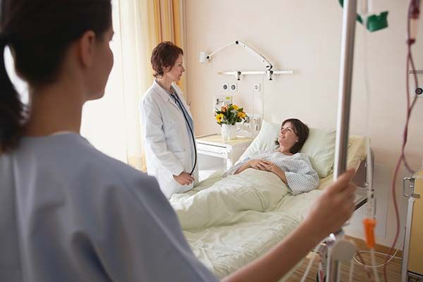 A doctor talks to a patient in a hospital bed while a nurse stands in the foreground, holding an IV pole.