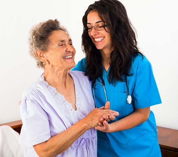 A healthcare worker in blue scrubs smiles while gently holding the hands of a person wearing a light purple top.