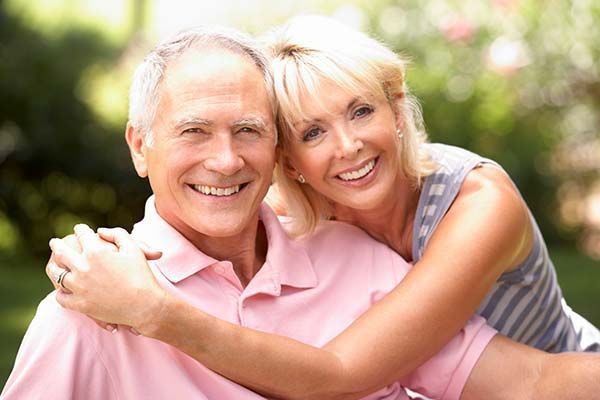 An older couple embraces and smiles at the camera while outdoors in a park setting.