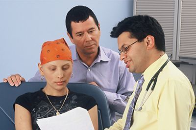 A doctor in a yellow shirt and stethoscope discusses medical documents with a patient wearing a headscarf and a companion.