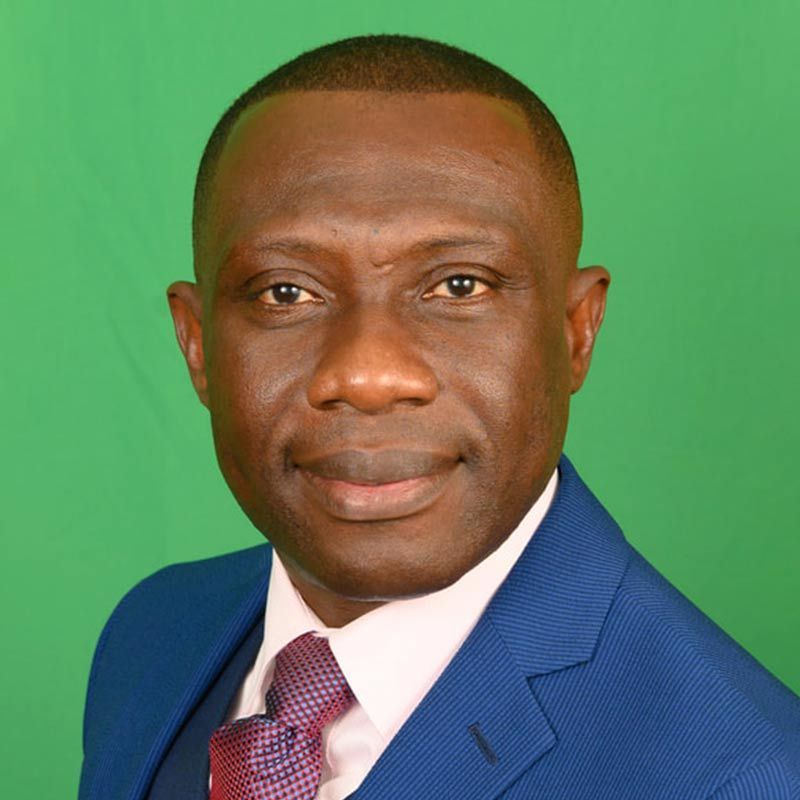 A man in a blue suit, white shirt, and patterned tie smiling against a plain green background.