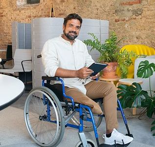 A person in a wheelchair smiles while holding a tablet in a bright office with plants and brick walls.