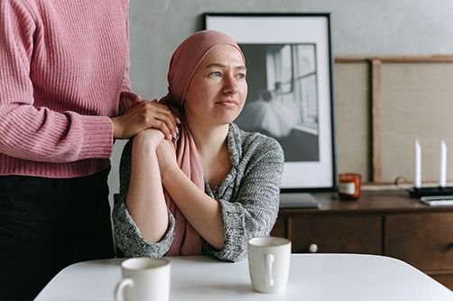 A person in a pink headscarf sits at a table with a companion, who rests a hand on their shoulder in a supportive gesture.
