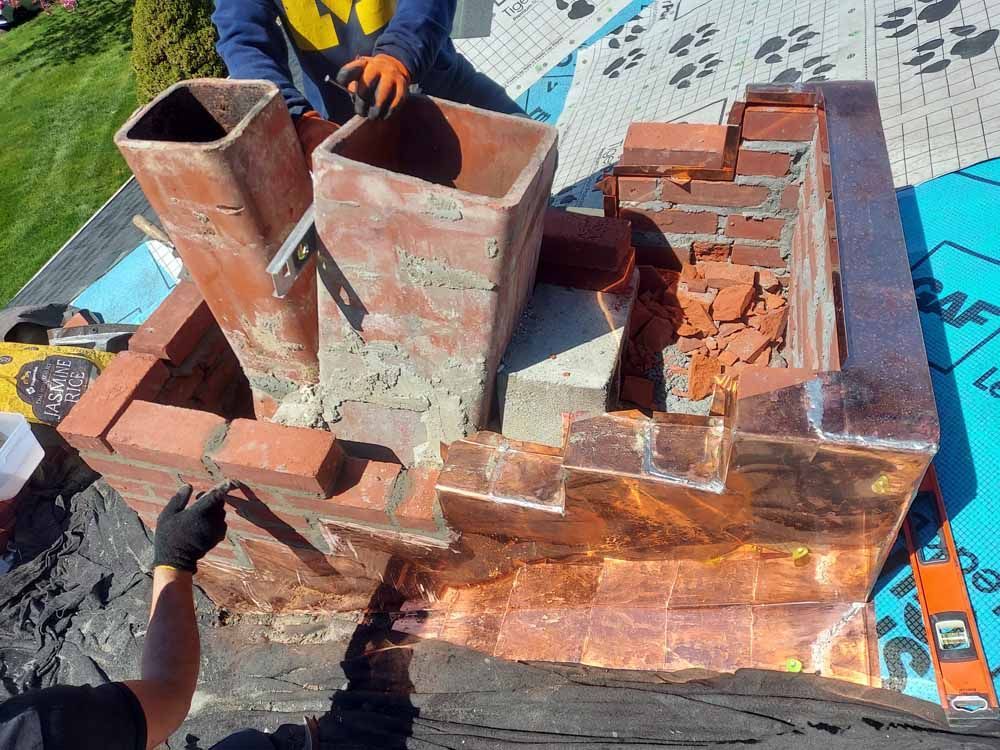 A man is working on a brick chimney on a roof.