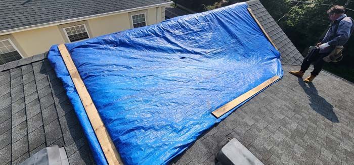 A man is standing on top of a roof with a blue tarp on it.