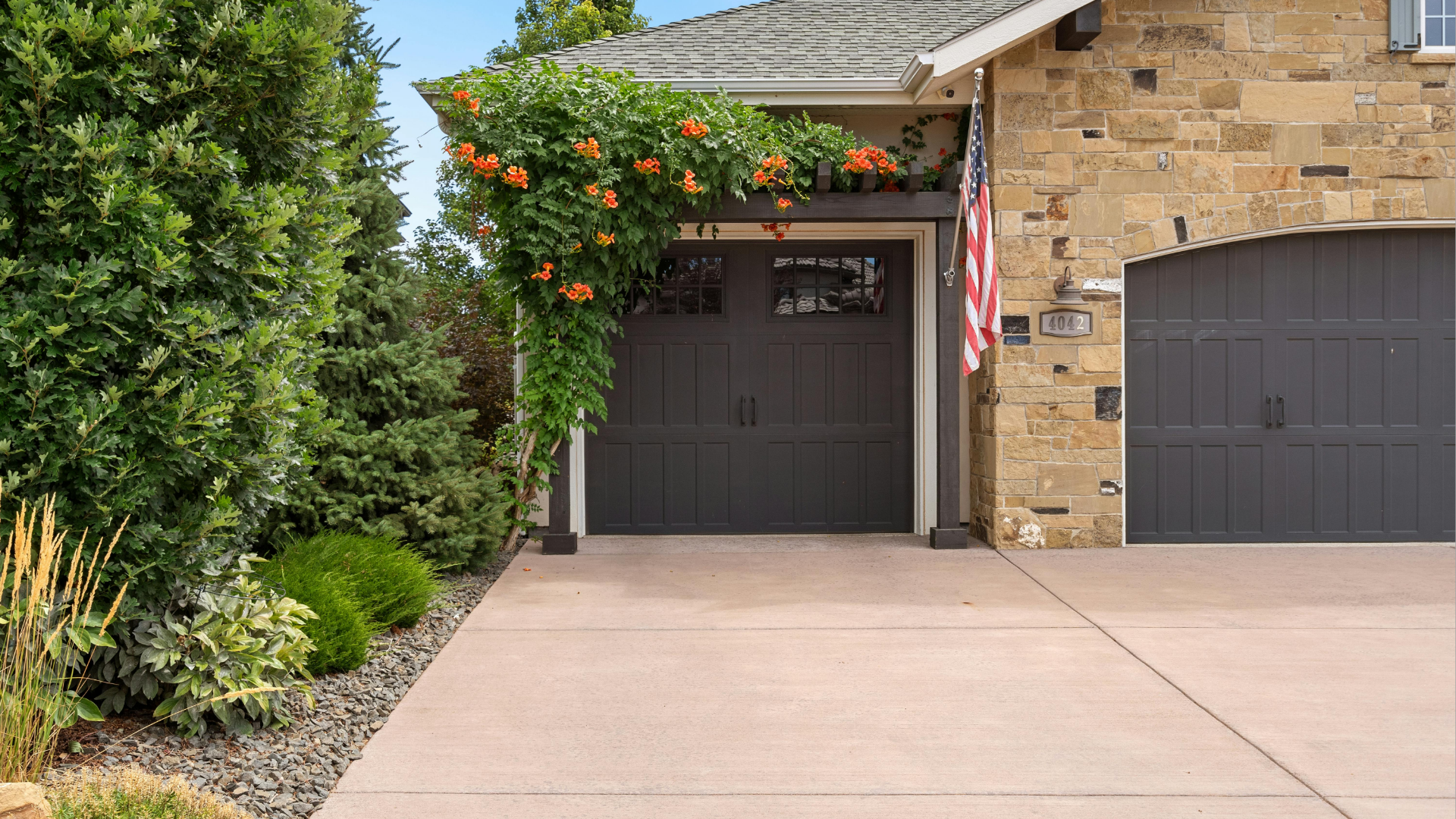 Grey garage doors with orange flowers and an American flag in front of a brick house.