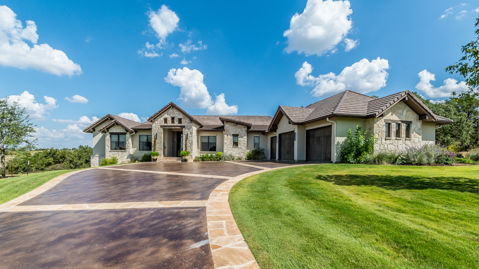 Sprawling stone house with long driveway, set against a blue sky with puffy clouds.