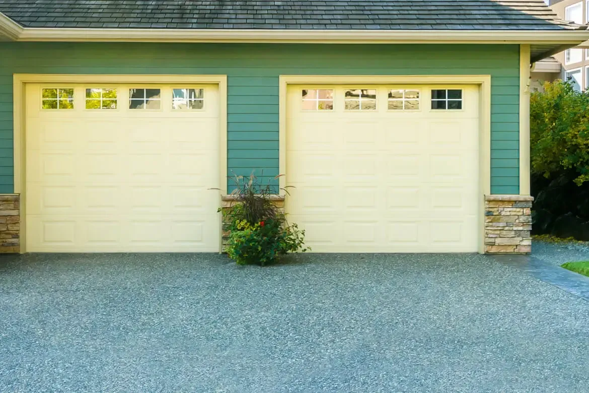 Two beige garage doors with glass windows, teal siding, gravel driveway.