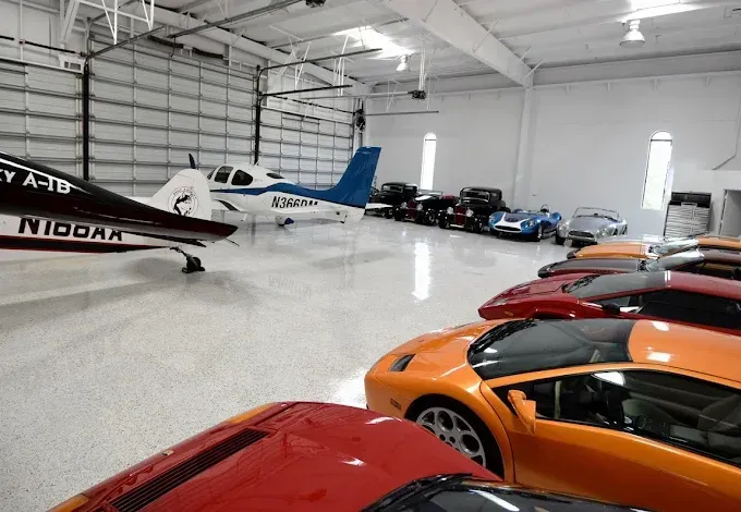 A hangar displays a small airplane and multiple exotic cars. Interior with white walls and floor.