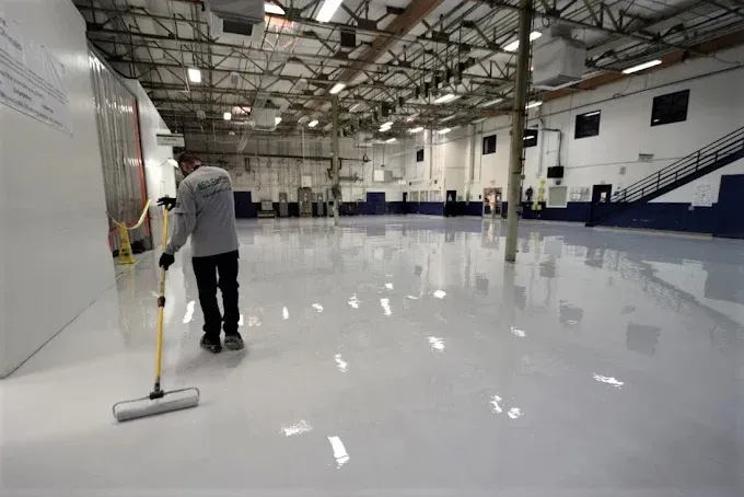 Person mopping a shiny, white epoxy floor in a large industrial warehouse.