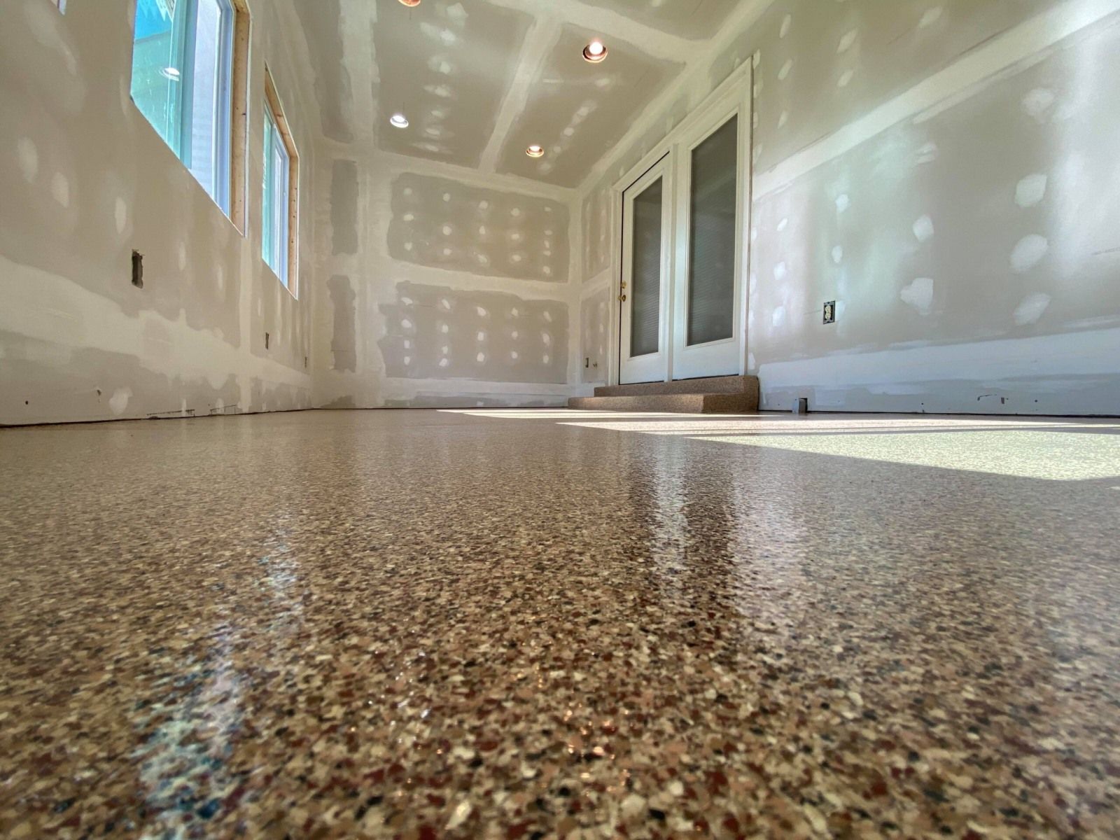 Room with a shiny, speckled floor. Walls and ceiling are unfinished drywall. Natural light streams through a window.