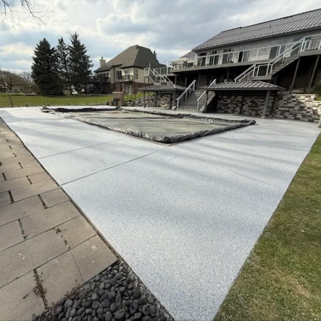 Concrete pool deck with a covered pool, next to a stone patio and grassy lawn. Cloudy sky.