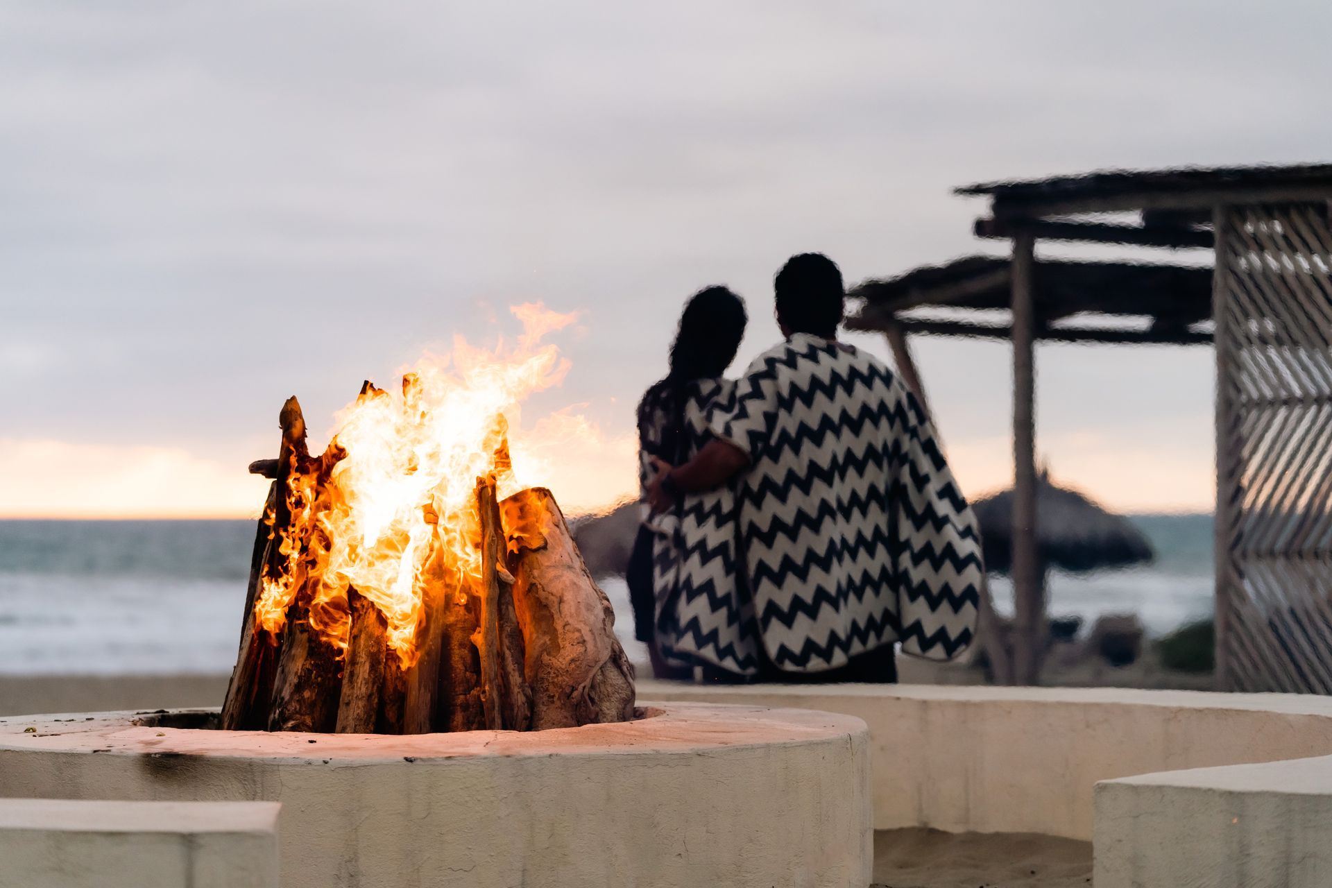 Couple embracing, watching fire pit on beach at dusk.