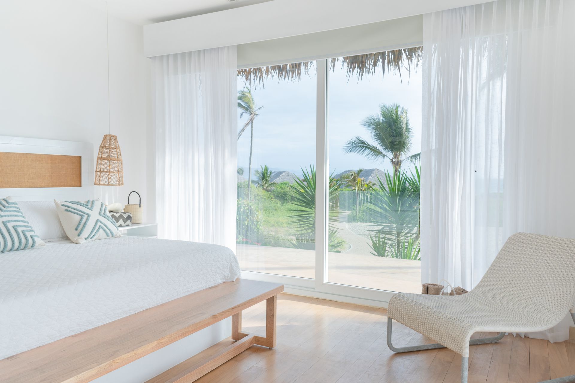 White bedroom with large window overlooking palm trees and beach; bed, bench, and chair visible.