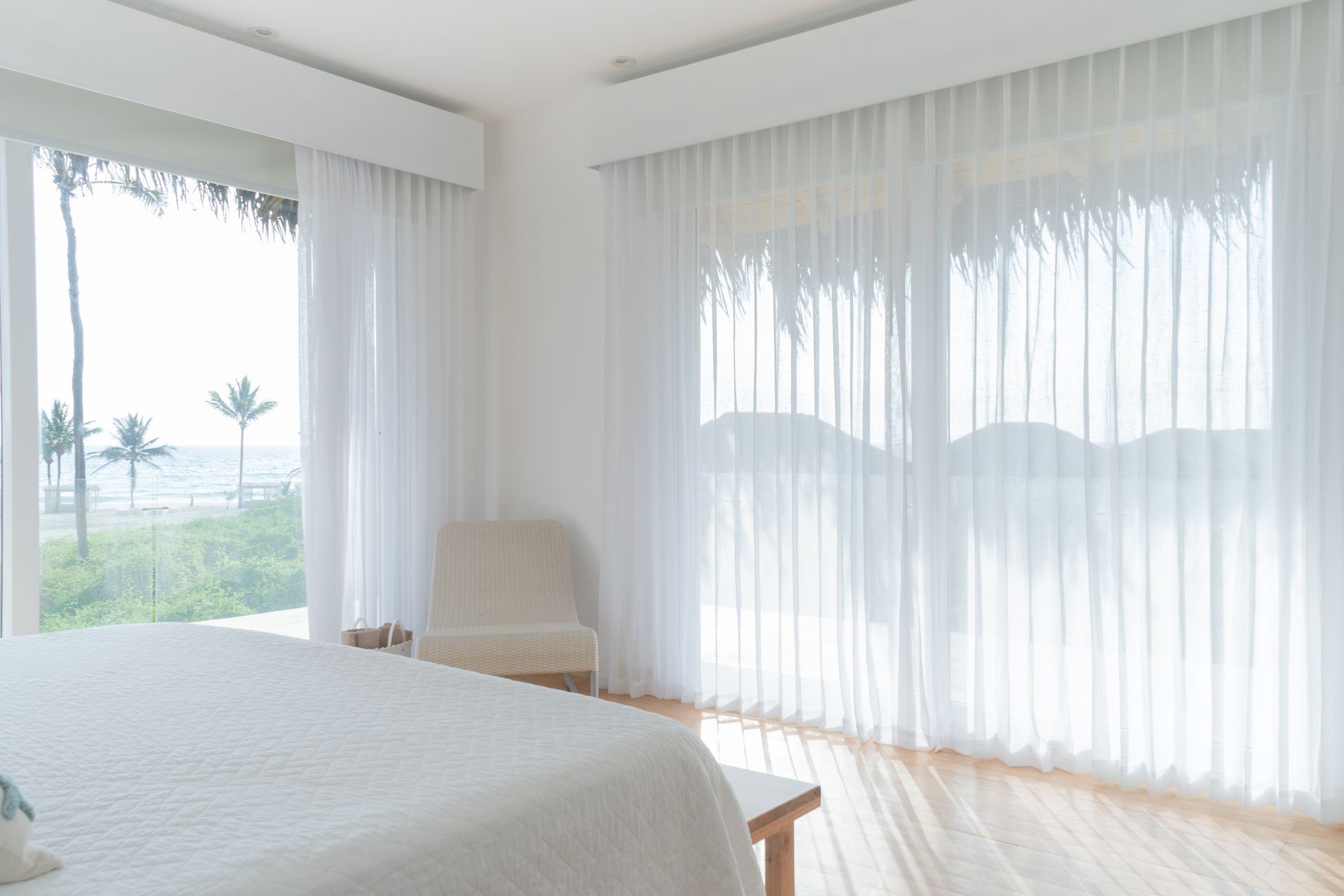 Bright bedroom with sheer white curtains, a bed, and a view of the ocean.