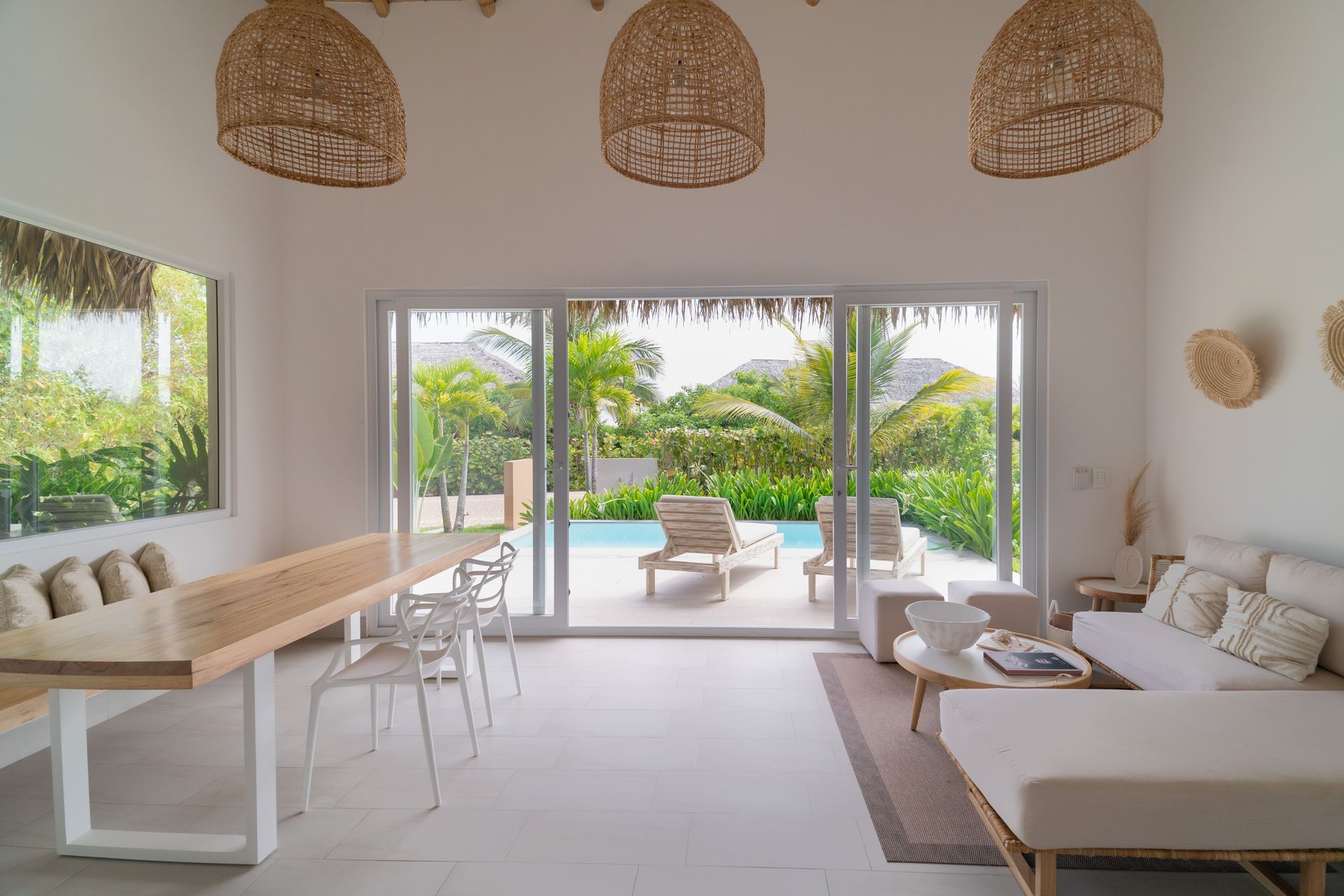 Bright living space with pool view; white walls, wooden dining table, sofa, woven light fixtures.