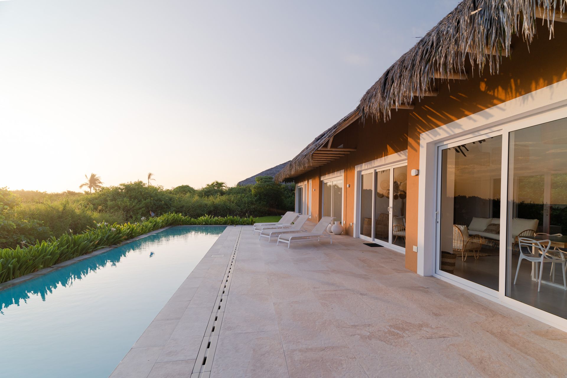 A tropical villa with a pool, sliding glass doors, and a thatched roof under a golden sunset.
