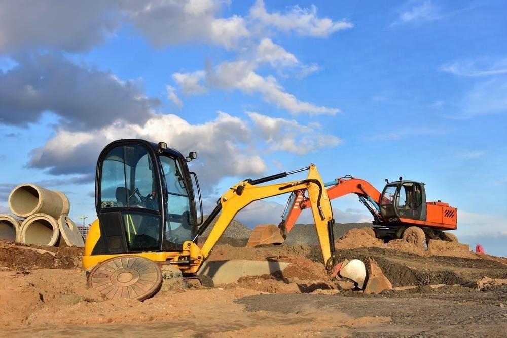 A yellow crawler excavator with a large scoop bucket is digging up dirt and rocks on a construction site, preparing the land for building a new house.