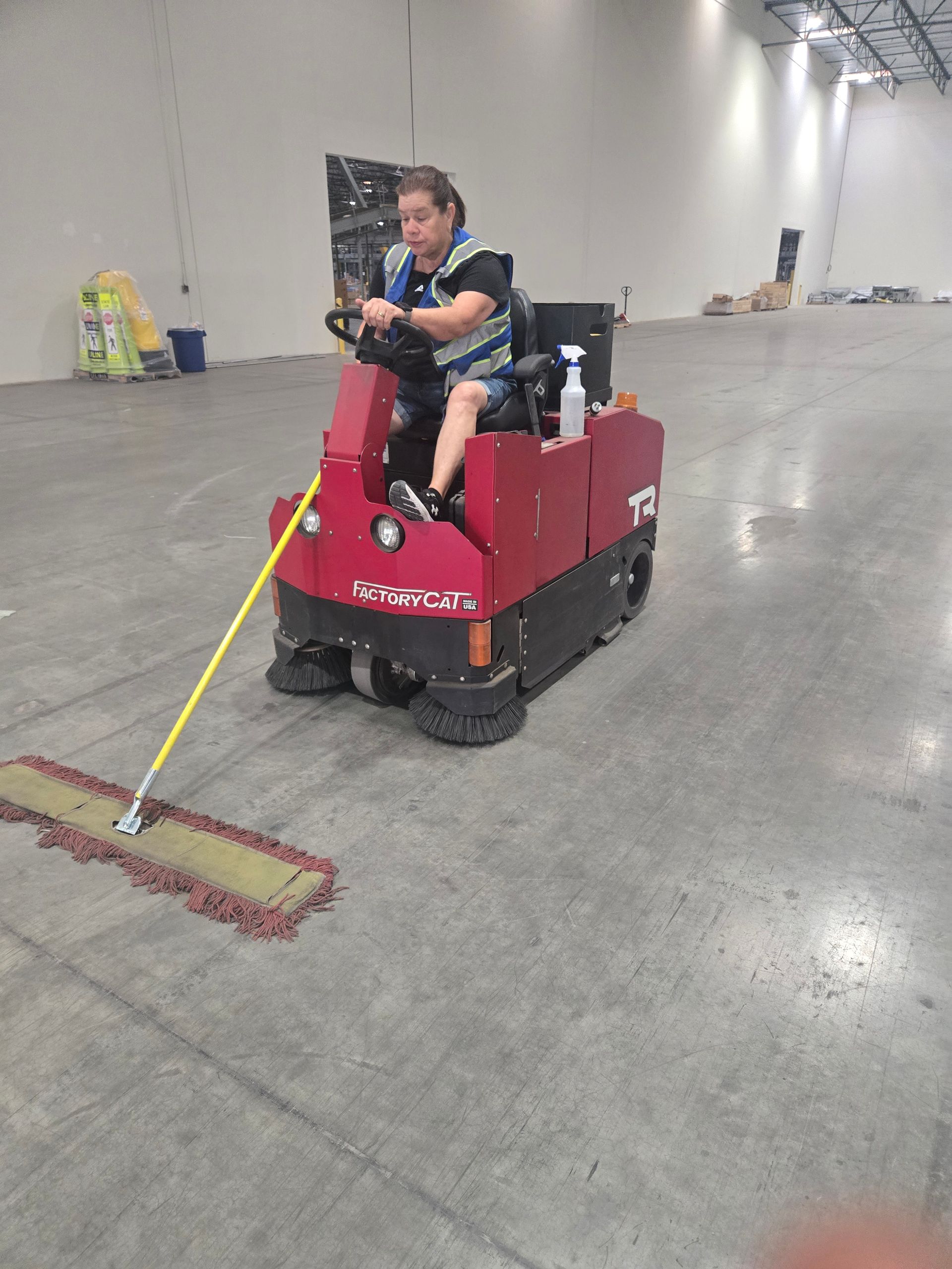 A person operates a red industrial floor cleaner while pushing a yellow mop in a large, empty warehouse.