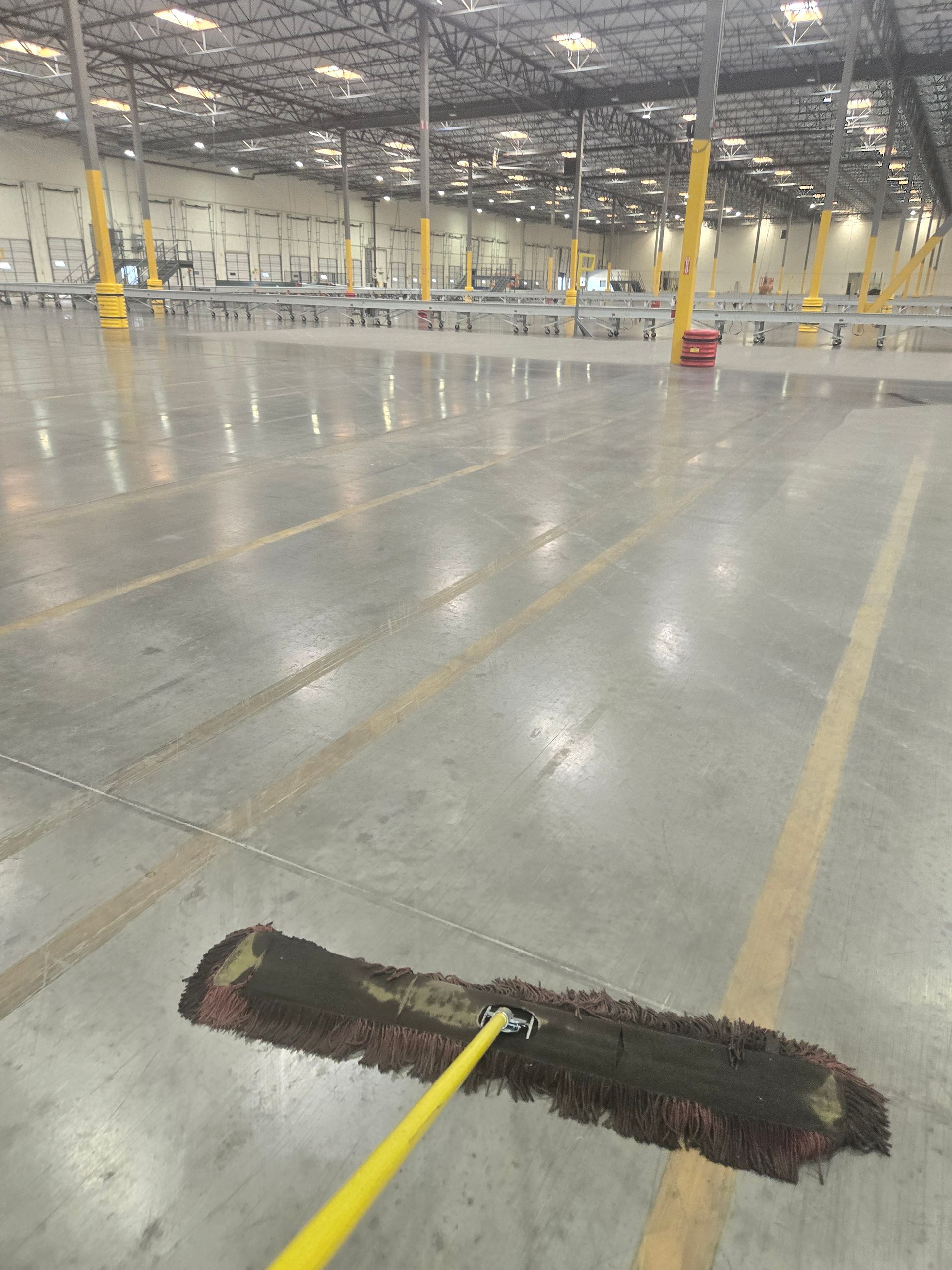 A janitorial dust mop rests on the polished concrete floor of a vast, empty warehouse with yellow support pillars.