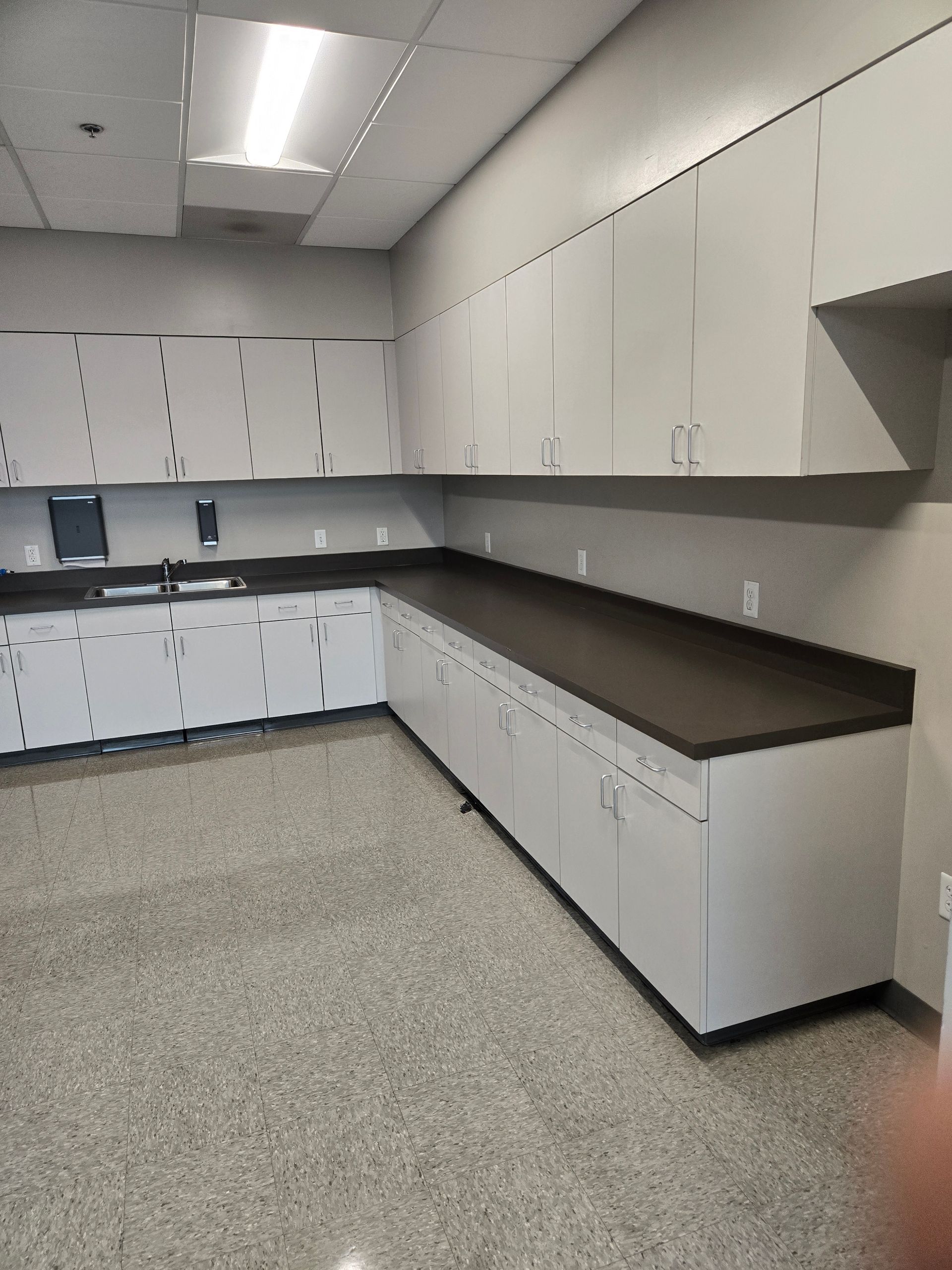 A breakroom with white cabinets, a dark countertop, a stainless steel sink, and speckled light-colored flooring.