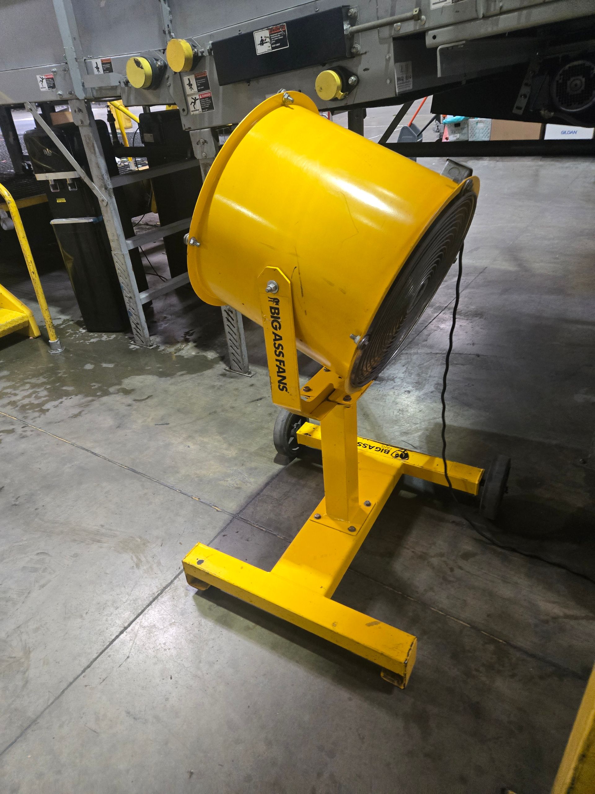 A bright yellow industrial floor fan with a circular cage, mounted on a mobile stand in a warehouse setting.