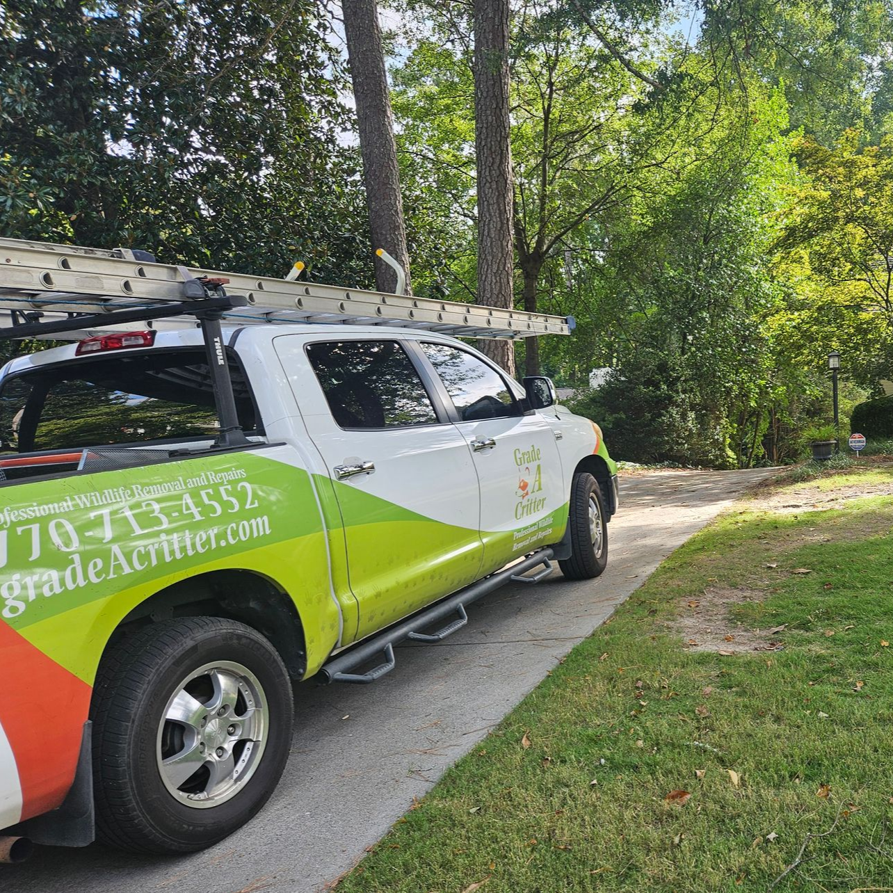 A side view of a white service truck parked on a driveway, featuring green and orange branding with a phone number.